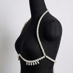 Peut inclure: Une cha&icirc;ne de corps en perles blanches drap&eacute;e sur un mannequin noir. La cha&icirc;ne comprend un collier, des bretelles et un design de poitrine inf&eacute;rieur avec des perles en forme de goutte suspendues. Les perles sont enfil&eacute;es ensemble, cr&eacute;ant un accessoire d&eacute;licat et &eacute;l&eacute;gant.
