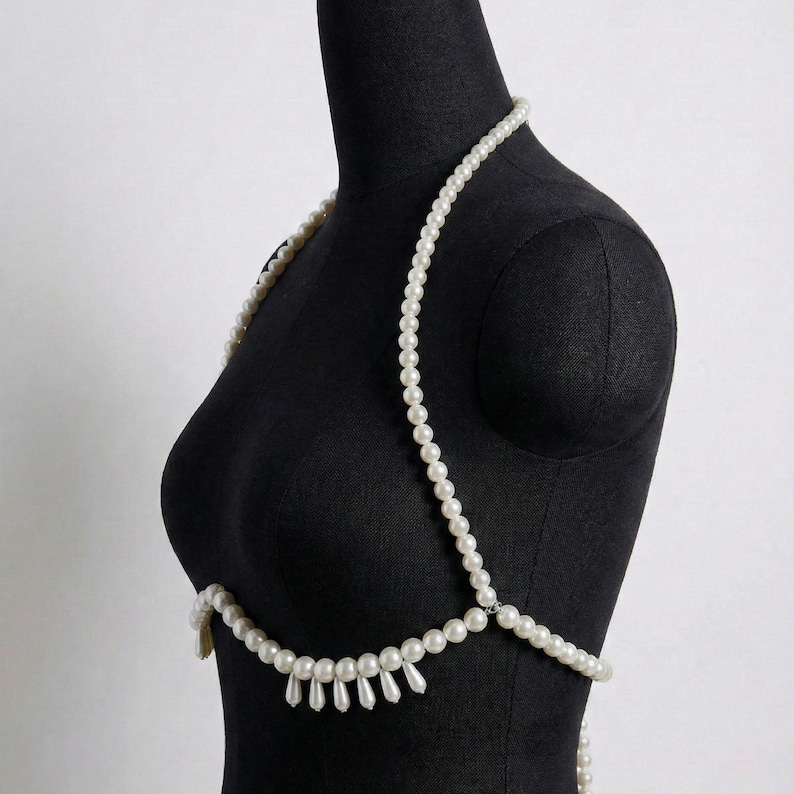 Peut inclure: Une cha&icirc;ne de corps en perles blanches drap&eacute;e sur un mannequin noir. La cha&icirc;ne comprend un collier, des bretelles et un design de poitrine inf&eacute;rieur avec des perles en forme de goutte suspendues. Les perles sont enfil&eacute;es ensemble, cr&eacute;ant un accessoire d&eacute;licat et &eacute;l&eacute;gant.