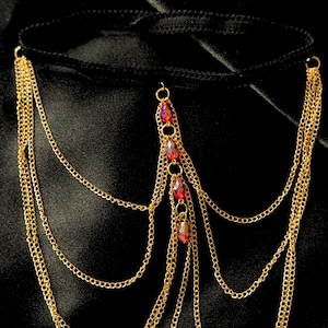 May include: Gold-colored body chain with multiple strands and red jewel accents. The chain features a black band and is designed to be worn around the waist or hips. The jewelry is displayed against a black background.