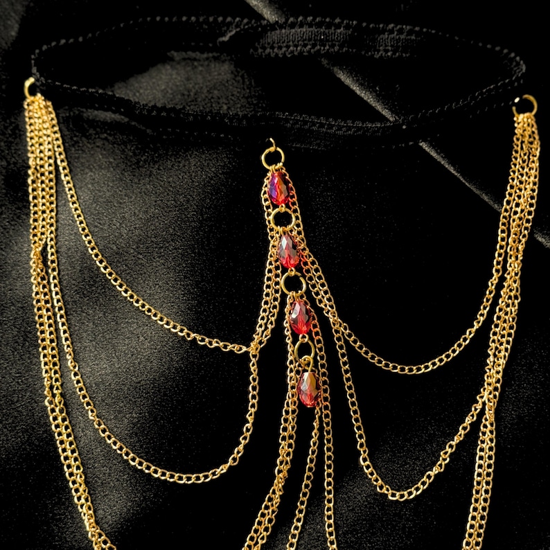 May include: Gold-colored body chain with multiple strands and red jewel accents. The chain features a black band and is designed to be worn around the waist or hips. The jewelry is displayed against a black background.