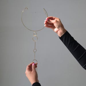 K&ouml;nnte beinhalten: Eine goldfarbene Halskette mit kreisf&ouml;rmigem Kragen und einem vertikalen Anh&auml;nger aus verbundenen Kreisen und Linien. Die Halskette wird vor einem grauen Hintergrund gehalten, um ihr zartes Design und ihre moderne &Auml;sthetik hervorzuheben.