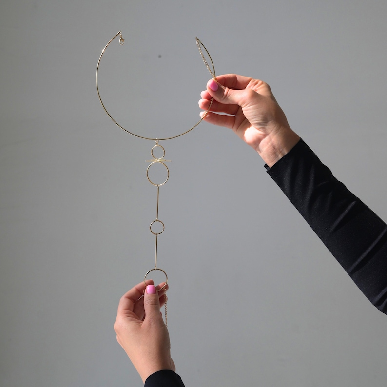 K&ouml;nnte beinhalten: Eine goldfarbene Halskette mit kreisf&ouml;rmigem Kragen und einem vertikalen Anh&auml;nger aus verbundenen Kreisen und Linien. Die Halskette wird vor einem grauen Hintergrund gehalten, um ihr zartes Design und ihre moderne &Auml;sthetik hervorzuheben.