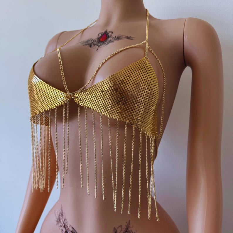 May include: Gold-colored chainmail top with a halter neck and fringe detailing. The top features a mesh design across the bust and is adorned with dangling chains. This piece is a statement accessory.