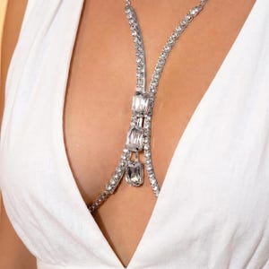 K&ouml;nnte beinhalten: Eine silberfarbene K&ouml;rperkette mit klaren, rechteckigen und runden Kristallen. Die Kette verl&auml;uft von den Schultern &uuml;ber die Brust. Der Schmuck wird &uuml;ber einem wei&szlig;en Oberteil getragen.