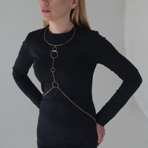 K&ouml;nnte beinhalten: Goldfarbene K&ouml;rperkette mit filigranem Design. Die Halskette hat einen oberen Teil im Choker-Stil, der durch eine Reihe von Kreisen und Linien verbunden ist, die &uuml;ber den Oberk&ouml;rper verlaufen. Eine Kette reicht bis zur Taille. Die Person tr&auml;gt ein schwarzes Kleid.