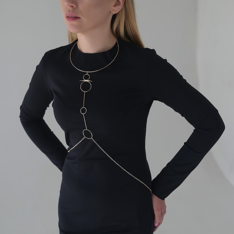 K&ouml;nnte beinhalten: Goldfarbene K&ouml;rperkette mit filigranem Design. Die Halskette hat einen oberen Teil im Choker-Stil, der durch eine Reihe von Kreisen und Linien verbunden ist, die &uuml;ber den Oberk&ouml;rper verlaufen. Eine Kette reicht bis zur Taille. Die Person tr&auml;gt ein schwarzes Kleid.