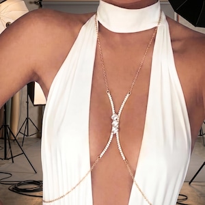 May include: A gold-toned body chain featuring a choker-style neck loop connected to a central vertical segment. This segment is embellished with a row of small clear crystals and three larger, faceted rectangular crystals arranged vertically.