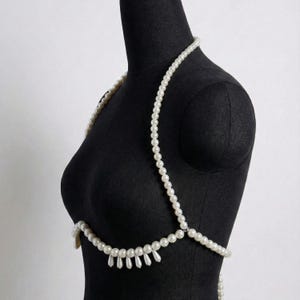 K&ouml;nnte beinhalten: Eine wei&szlig;e Perlen-K&ouml;rperkette drapiert auf einer schwarzen Schaufensterpuppe. Die Kette hat eine Halskette, Schultergurte und ein unteres Brustdesign mit h&auml;ngenden tropfenf&ouml;rmigen Perlen. Die Perlen sind aufgereiht und bilden ein zartes und elegantes Accessoire.