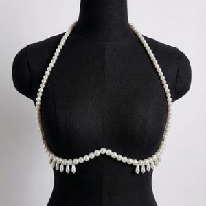 May include: A white pearl body chain necklace. The necklace is draped over a black mannequin. The necklace has a halter-style design with a series of small pearls and teardrop-shaped pearl pendants.