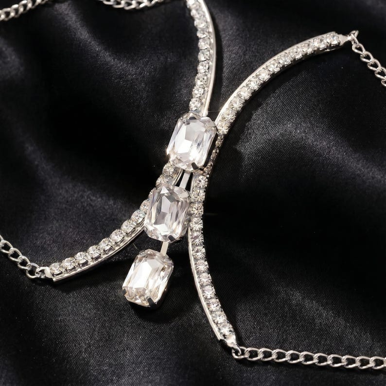 K&ouml;nnte beinhalten: Silberner K&ouml;rperschmuck mit drei gro&szlig;en, klaren rechteckigen Kristallen und kleineren, umgebenden klaren Kristallen. Der Schmuck hat eine Kette und ist vor einem schwarzen Satinhintergrund platziert.
