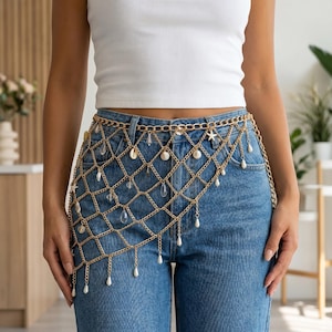May include: Gold-colored chain belt with a net design, adorned with seashells, clear crystals, and dangling faux pearls. The belt is worn over blue jeans, creating a stylish accessory. The belt is a fashion accessory.