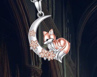 May include: A silver necklace with a crescent moon pendant featuring a small fox sitting on the moon with pink flowers. The fox is made of rose gold.