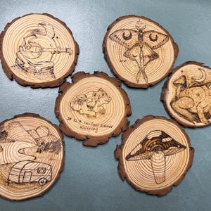 May include: Set of six wood slice coasters with pyrography designs. The designs include a musician, a moth, a frog, a mushroom, a camper, and a cow. The coasters are made of natural wood with a dark brown bark edge.