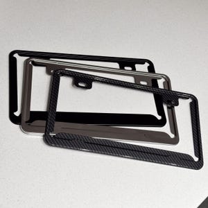 May include: Three car license plate frames in black, silver, and carbon fibre. The black frame is solid, the silver frame is metallic, and the carbon fibre frame has a textured weave pattern. All frames have a rectangular shape with rounded corners.