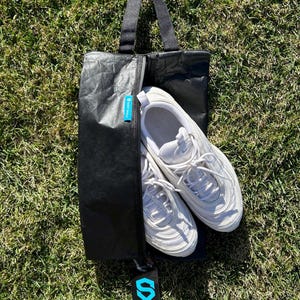 May include: Black shoe bag with a zipper closure and a black handle. The bag is made of a crinkled material and has a blue logo on the front. A pair of white sneakers are inside the bag.