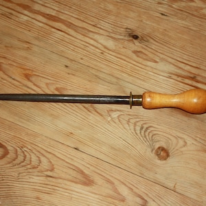 May include: A steel knife sharpening rod with a wooden handle and a metal ring. The rod is dark grey, and the handle is light brown. The sharpening rod is lying on a wooden surface.