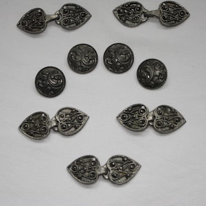 May include: A collection of silver-toned metal fasteners and buttons. There are five heart-shaped clasps featuring intricate filigree wire patterns and four circular buttons with embossed swirling scrollwork designs on their surfaces.