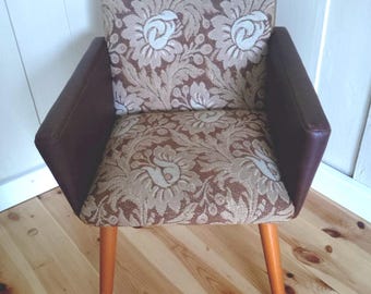 May include: A vintage armchair with a floral patterned seat and backrest. The chair features brown leather armrests and light brown wooden legs. The floral pattern includes white and beige flowers on a brown background.