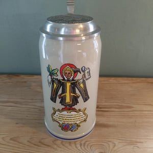 May include: A tall, cylindrical ceramic beer stein with a light beige finish and a metallic lid. The front features a colourful illustration of a figure in traditional robes holding a radish and a mug, with German text and a crest below.