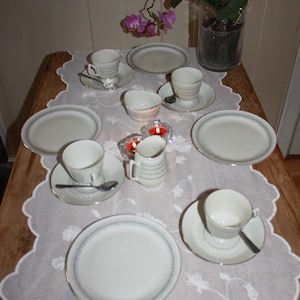 May include: A table setting featuring white teacups, saucers, and plates with a blue and gold trim. Small spoons and a creamer are also present. The table is covered with a white lace tablecloth, and a vase with an orchid is in the background.