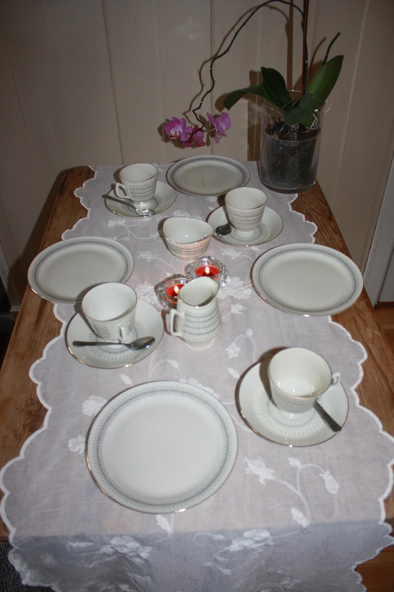 May include: A table setting featuring white teacups, saucers, and plates with a blue and gold trim. Small spoons and a creamer are also present. The table is covered with a white lace tablecloth, and a vase with an orchid is in the background.