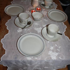 May include: A formal dining table setting with white porcelain dishes, cups, saucers, and a creamer. Each piece has a blue and gold trim. The table is covered with a white lace tablecloth, and two lit candles are visible.