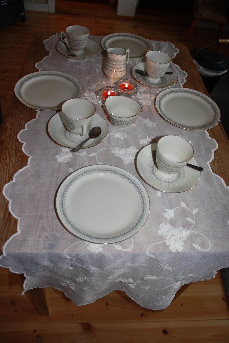 May include: A formal dining table setting with white porcelain dishes, cups, saucers, and a creamer. Each piece has a blue and gold trim. The table is covered with a white lace tablecloth, and two lit candles are visible.