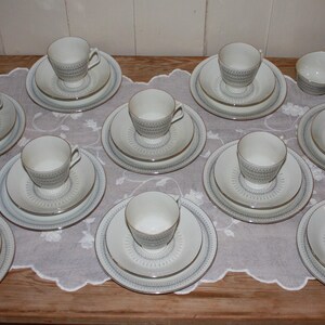 May include: A collection of white teacups, saucers, and plates arranged on a wooden table covered with a white lace tablecloth. The teacups have a textured design, and the plates have a gold trim. A small creamer and sugar bowl are also present.