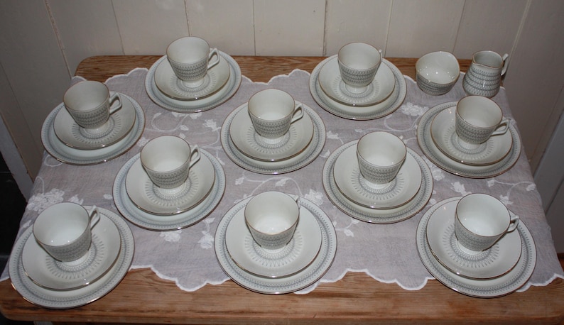 May include: A collection of white teacups, saucers, and plates arranged on a wooden table covered with a white lace tablecloth. The teacups have a textured design, and the plates have a gold trim. A small creamer and sugar bowl are also present.
