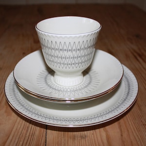 May include: A white teacup and saucer set with a gray geometric pattern and gold trim. The teacup sits on a matching saucer and a larger plate, creating a layered effect. The set is designed for serving tea or coffee.