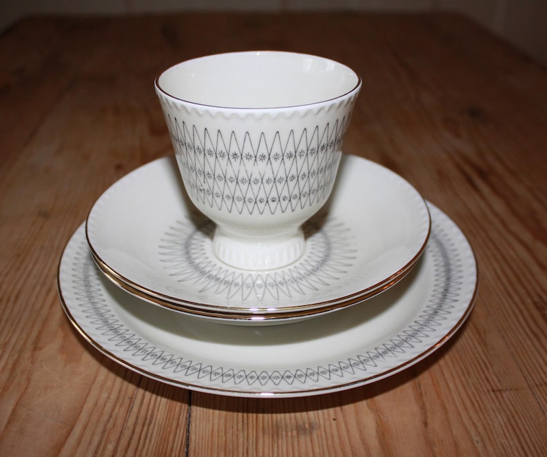 May include: A white teacup and saucer set with a gray geometric pattern and gold trim. The teacup sits on a matching saucer and a larger plate, creating a layered effect. The set is designed for serving tea or coffee.