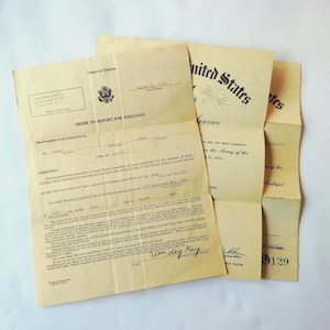 May include: Vintage military documents, including an "Order to Report for Induction" from the U.S. government. The papers are aged with visible creases and text, featuring the U.S. seal and text like "United States Service".