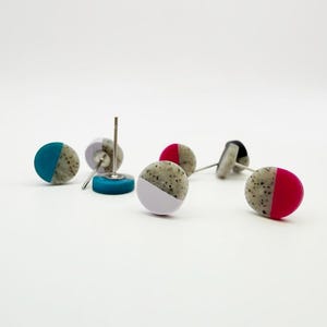 May include: A collection of colorful stud earrings. Each earring features a speckled, gray half-circle paired with a solid color half-circle in teal, lavender, red, and black. The earrings are displayed on a white surface.