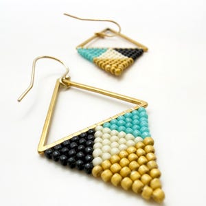 May include: A pair of triangular drop earrings featuring gold-tone metal frames and hook wires. The lower portion of each triangle is filled with a geometric pattern of small beads in turquoise, cream, black, and metallic gold colors.