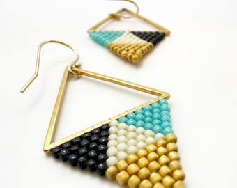 May include: A pair of triangular drop earrings featuring gold-tone metal frames and hook wires. The lower portion of each triangle is filled with a geometric pattern of small beads in turquoise, cream, black, and metallic gold colors.