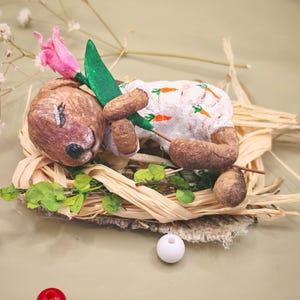 May include: A small brown rabbit figurine lying on a bed of raffia, wearing a white onesie with an orange carrot print. The rabbit holds a pink tulip with a green stem and leaves. The figurine is surrounded by small green leaves and white beads.