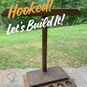 May include: A wooden hook and ring toss game with the text "Hooked! Let's Build It!". The game has a dark brown finish, metal rings, and a base with circular cutouts. Perfect for outdoor fun.