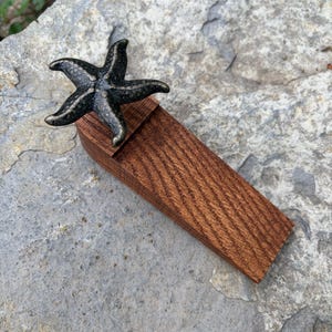 May include: A decorative doorstop featuring a bronze-colored starfish atop a rectangular, reddish-brown wooden base. The doorstop is designed to hold a door open and adds a nautical touch to any room.