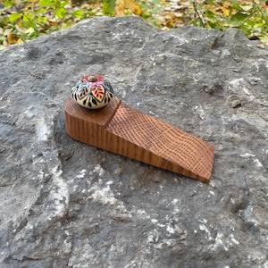 Pode incluir: Um calço de porta de madeira com um puxador decorativo de cerâmica pintado à mão. O puxador apresenta um design floral com detalhes em vermelho, rosa, azul e preto. O calço de porta é feito de madeira castanha clara e é angulado.