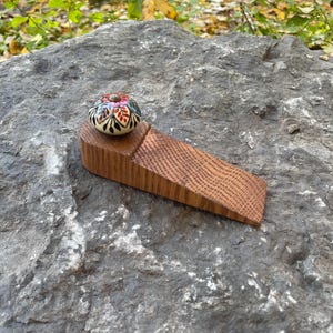 May include: A wooden doorstop with a decorative, hand-painted ceramic knob. The knob features a floral design with red, pink, blue, and black accents. The doorstop is made of light brown wood and is angled for functionality.