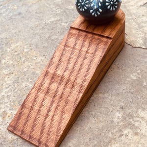 May include: A wooden doorstop with a decorative, dark blue and white floral-patterned knob. The doorstop is made of stained wood with a diagonal grain pattern. The knob has a gold-colored top.