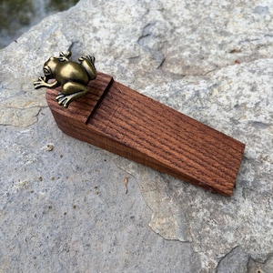 May include: A wooden doorstop with a bronze frog figurine on top. The doorstop is rectangular and made of dark brown wood. The frog is detailed with visible eyes and legs. The doorstop is resting on a gray stone surface.