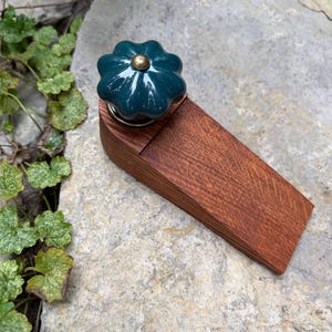 May include: A doorstop featuring a wedge-shaped wooden base with a visible grain pattern and a dark teal, flower-shaped ceramic knob attached to the top. The knob has a small, centered brass-colored metal accent at its highest point.