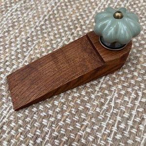 Może przedstawiać: Drewniany ogranicznik do drzwi z jasnozielonym, kwiatowym pokrętłem i mosiężnym środkiem. Ogranicznik do drzwi wykonany jest z ciemnobrązowego drewna i ma kształt klina. Pokrętło jest przymocowane do małej, okrągłej metalowej podstawy.