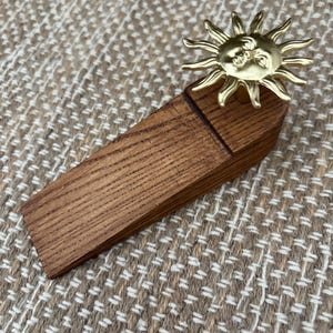 Puede incluir: Un tope de puerta de madera con un diseño de sol de latón. El tope de puerta está hecho de madera marrón oscuro y tiene forma de cuña. El sol tiene una cara en el centro y rayos que se extienden hacia afuera. El tope está diseñado para mantener una puerta abierta.