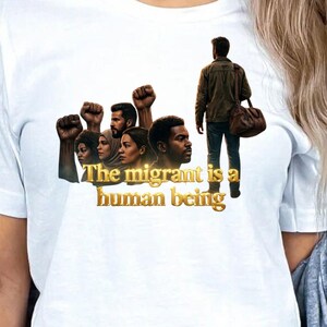 Peut inclure: T-shirt blanc avec un graphisme de visages divers, des poings levés et une personne s'éloignant avec un sac. Le texte "The migrant is a human being" est en lettres dorées. Un t-shirt de déclaration.