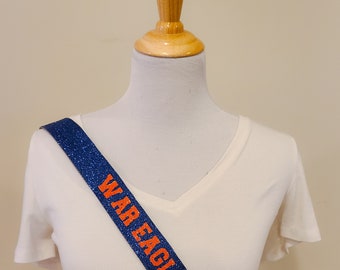 Op de afbeelding: Een blauwe glitter sjerp met de woorden "WAR EAGLE" in oranje letters. De sjerp is gedrapeerd over een wit v-hals T-shirt op een mannequin. De sjerp is een decoratief accessoire.