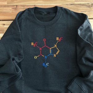 May include: A dark gray sweatshirt with a colorful embroidered chemical structure of caffeine on the front. The structure is made up of a six-sided ring with two nitrogen atoms, two oxygen atoms, and three carbon atoms. The carbon atoms are labeled with the chemical formula "H3C".