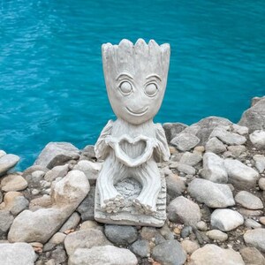 May include: A white concrete statue of a character holding a heart in its hands. The statue is sitting on a bed of small rocks near a body of water.