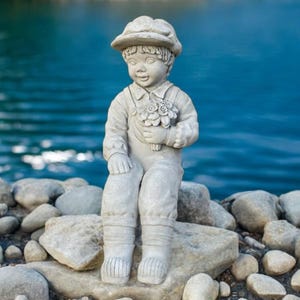 May include: A concrete statue of a young child wearing a hat and overalls, holding a bouquet of flowers. The statue is seated on a large rock, with a body of water in the background. The statue is a light gray color.
